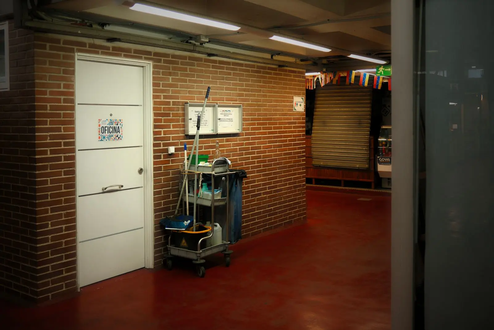 Schoonmaakwagen met bezem, mop en flessen in een lege gang tegen een bakstenen muur, naast een kantoordeur