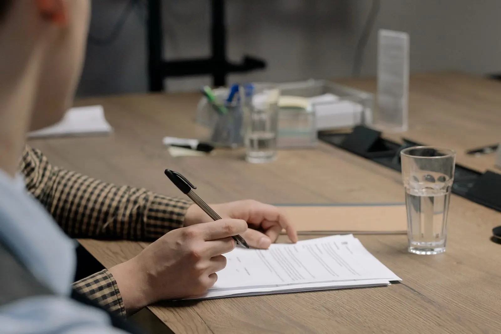 Iemand met een pen in de hand buigt zich over een document aan een houten vergadertafel met glazen water