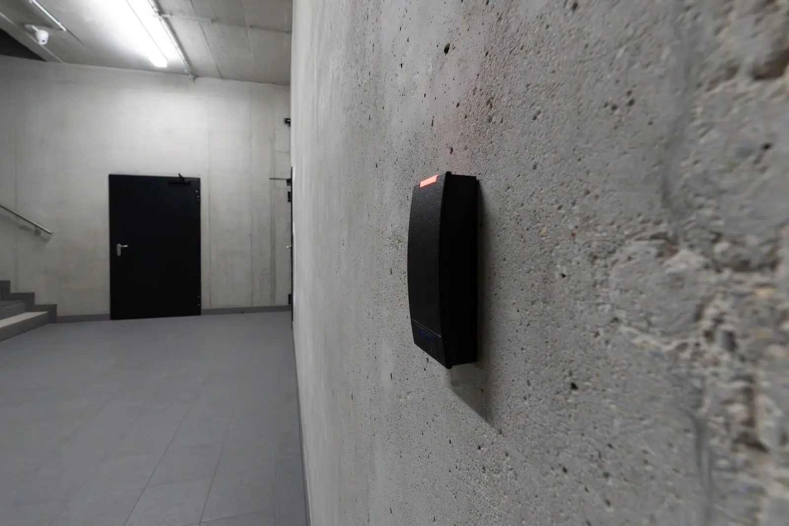 Zwarte pasjeslezer voor toegangscontrole aan een betonnen muur, met een gang en een deur op de achtergrond