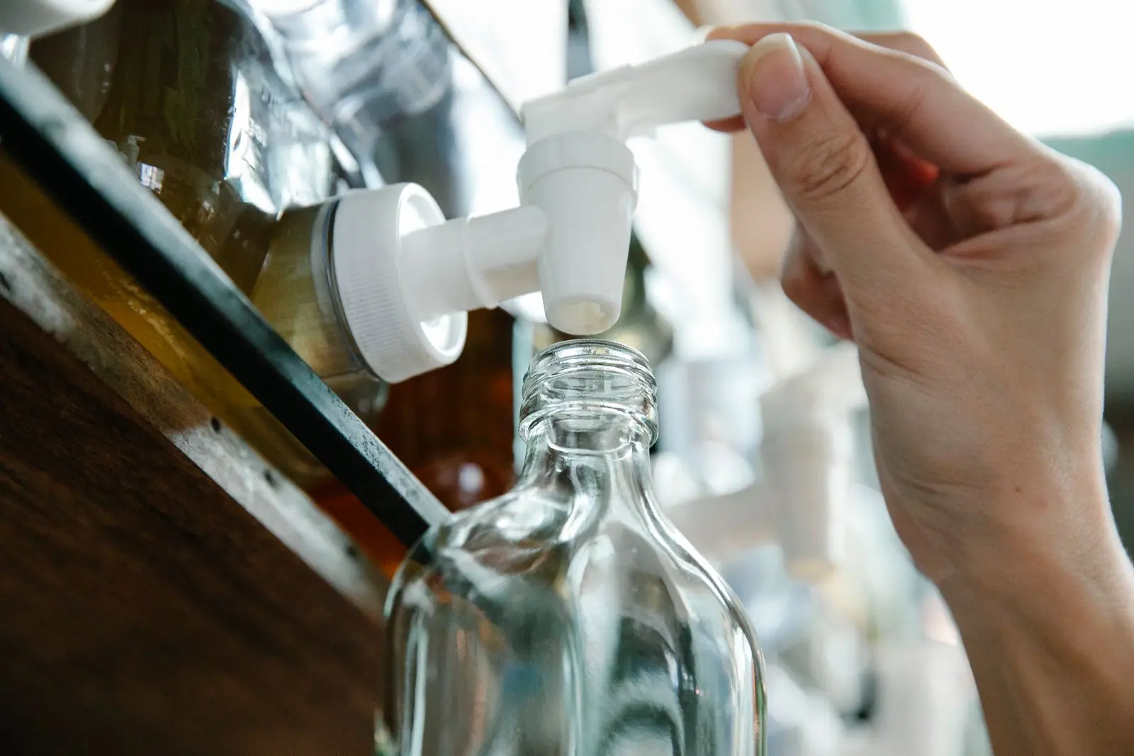 Hand opent het kraantje van een grote voorraaddispenser om een lege glazen fles bij te vullen
