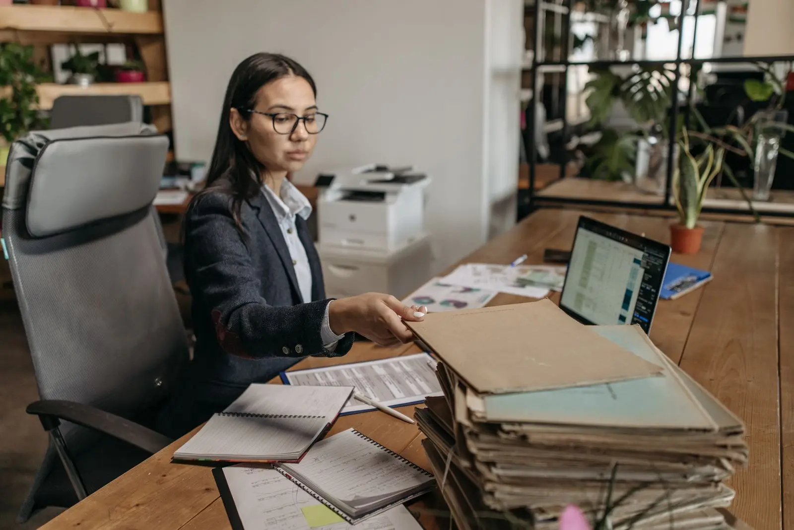 Vrouw aan een bureau met een stapel papieren dossiermappen naast een laptop met een spreadsheet