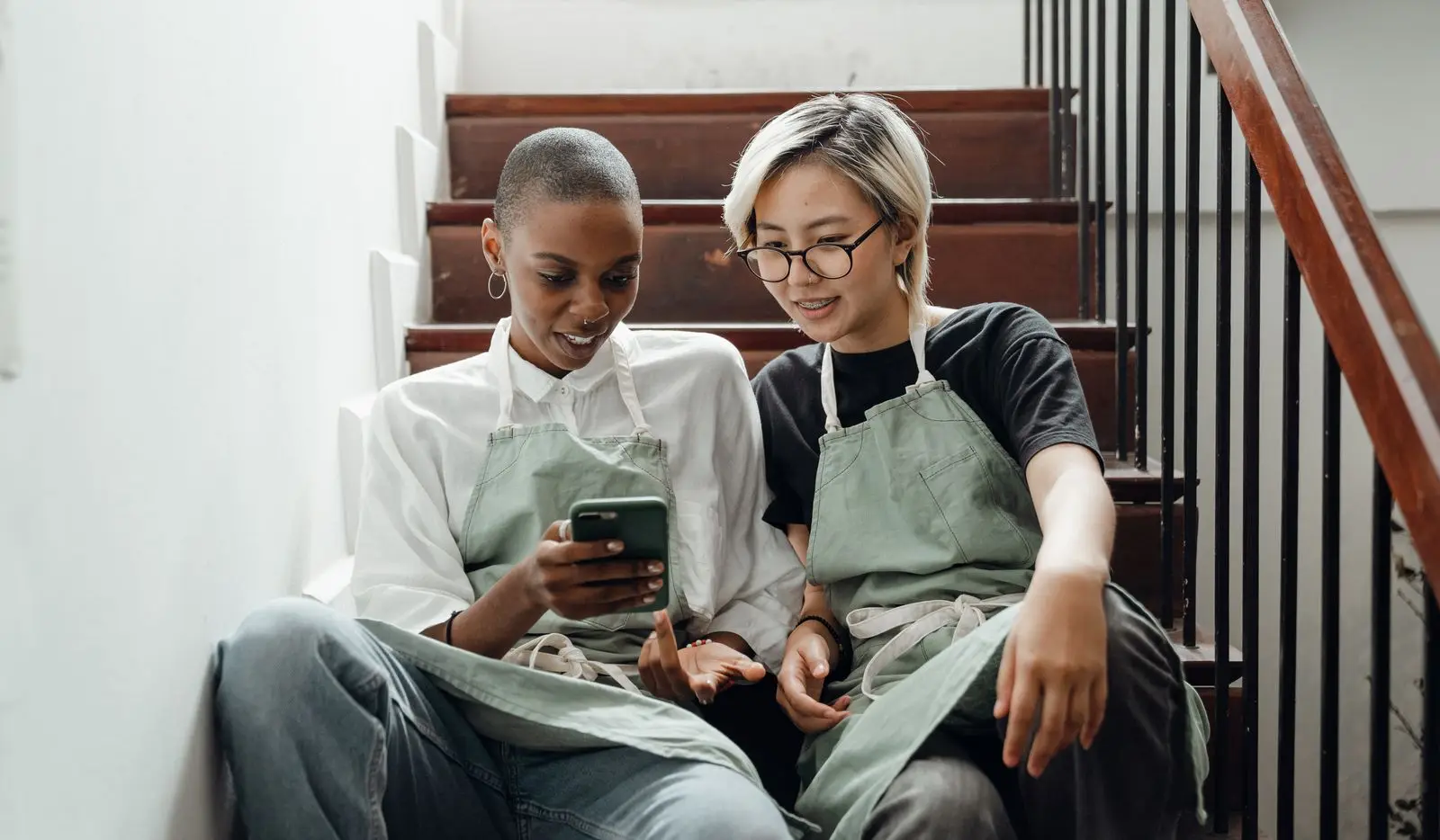 Twee jonge vrouwen in groene werkschorten zitten naast elkaar op een trap en kijken samen naar het scherm van een smartphone