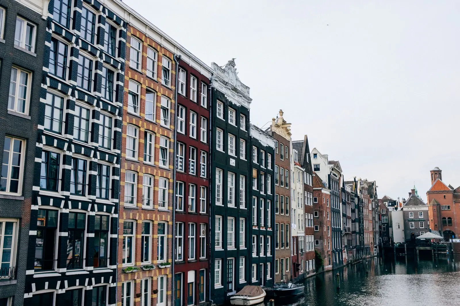 Rij smalle grachtenpanden in Amsterdam met bakstenen gevels en hoge ramen langs een gracht met twee afgemeerde bootjes