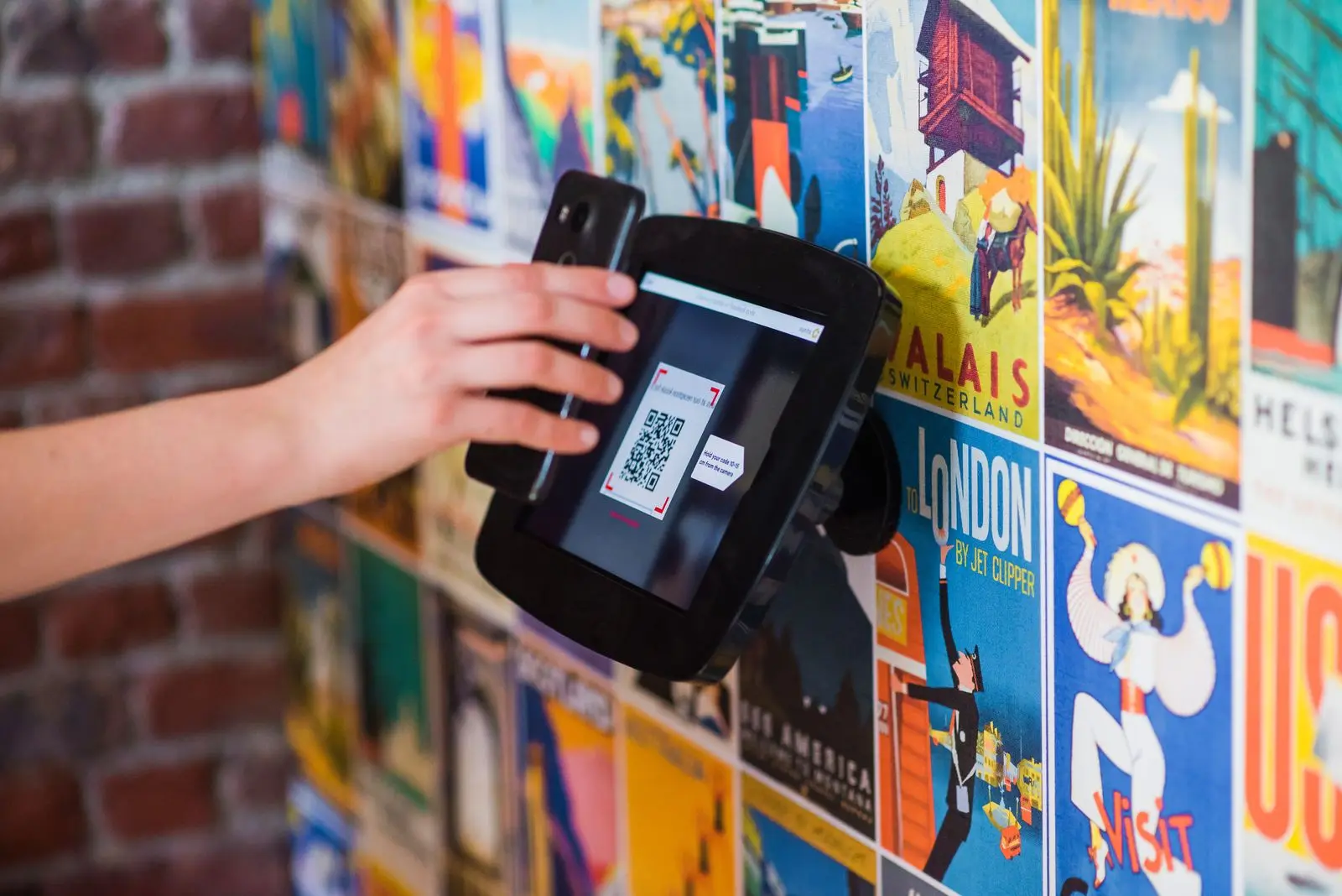 Hand houdt een smartphone voor een scanner aan de muur die een QR-code in beeld heeft, tegen een wand met kleurige reisposters