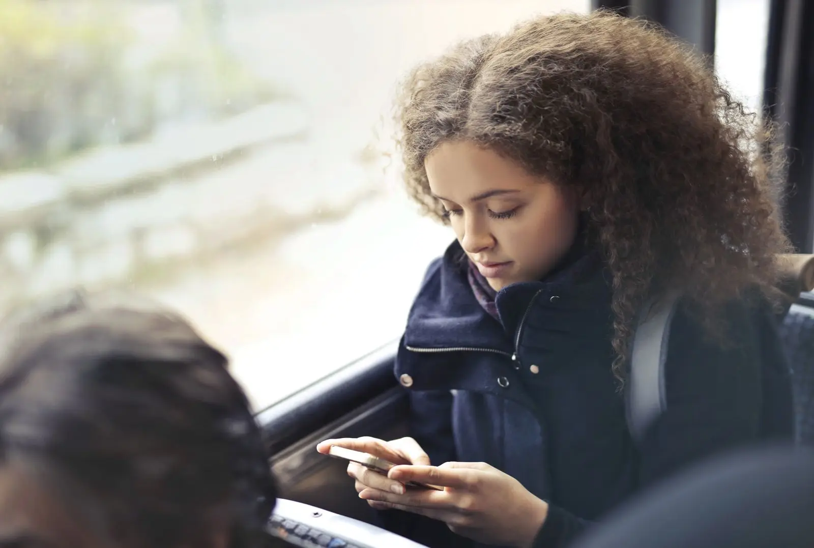 Jonge vrouw met krullend haar zit in de bus bij het raam en typt iets op haar smartphone.