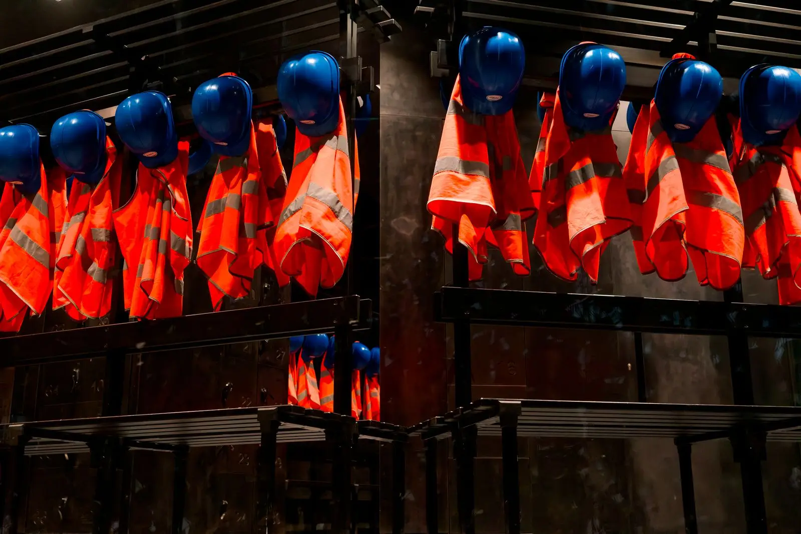 Rijen oranje veiligheidshesjes met blauwe bouwhelmen erboven hangen aan haken in een donkere ruimte.