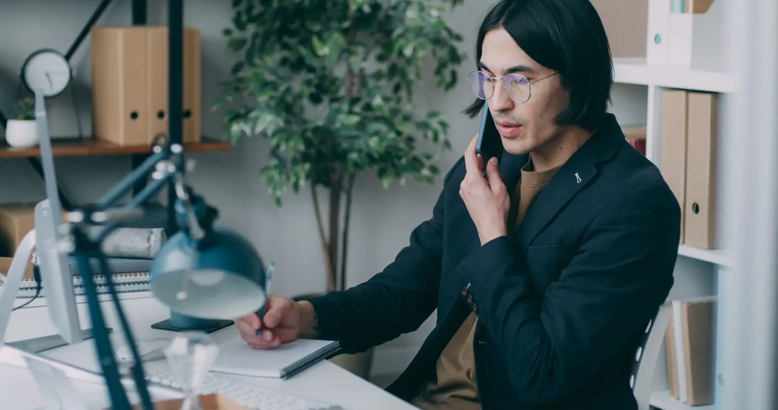 Man met bril belt aan zijn bureau en maakt aantekeningen in een notitieblok, met een plant en ordners achter hem.