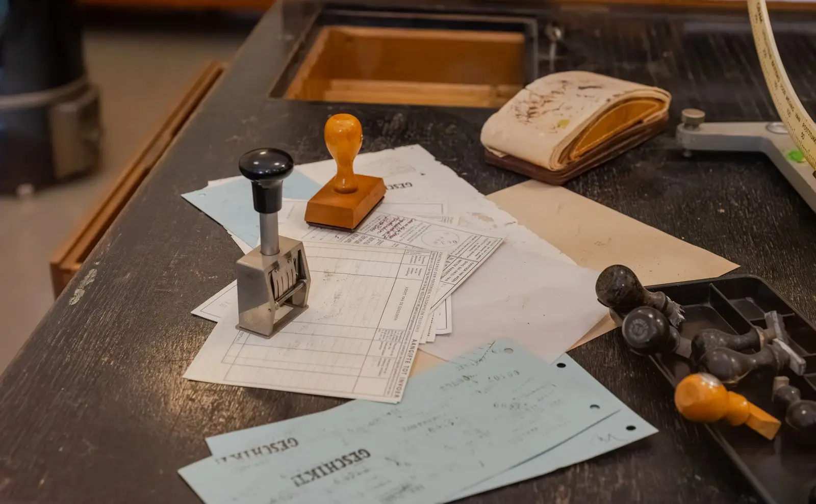 Oude stempels en ingevulde formulieren op een houten bureau; op twee blauwe strookjes staat 'GESCHIKT!' gestempeld.