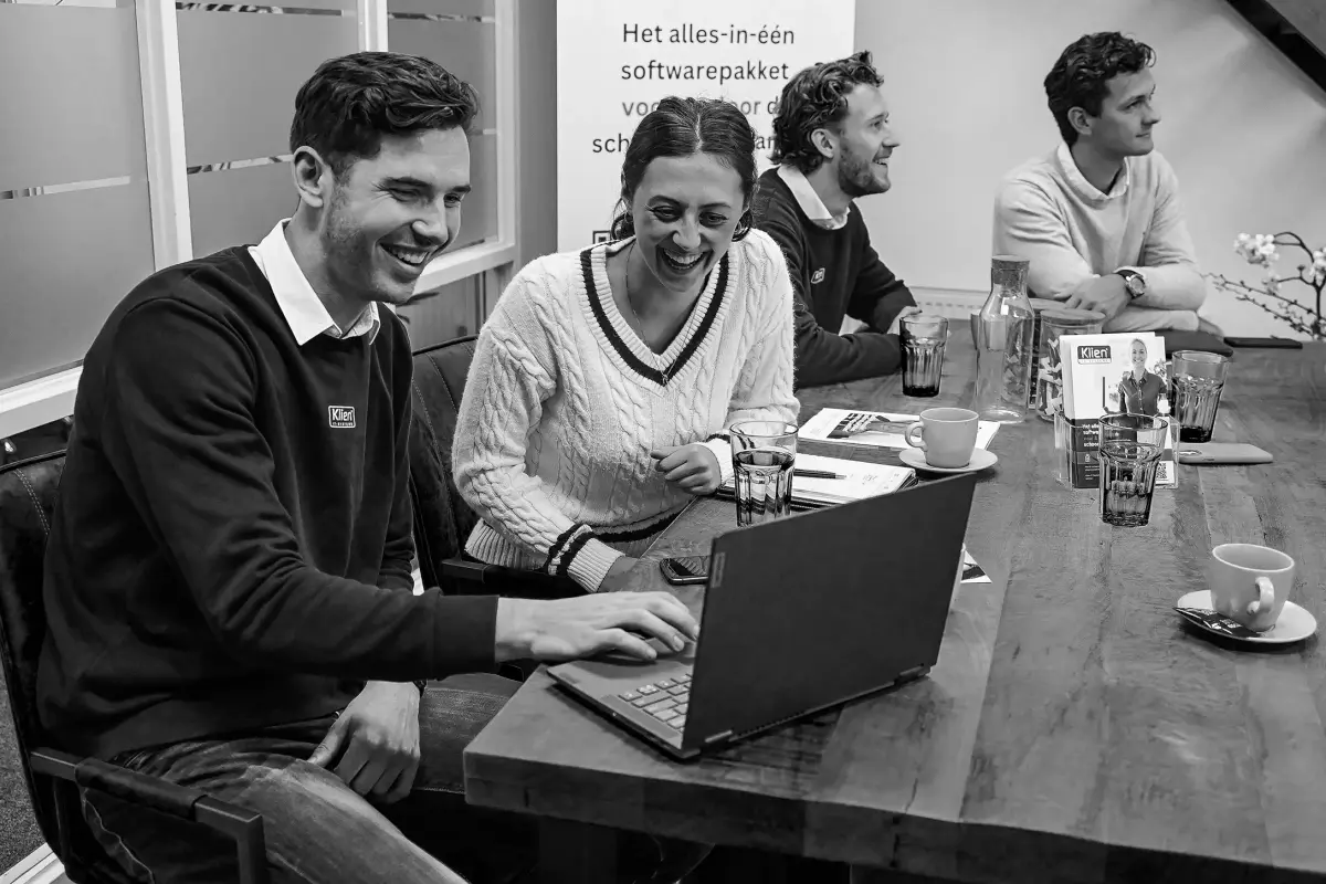 Zwart-witfoto van medewerkers van Klien-IT aan een vergadertafel, twee van hen lachend achter een laptop