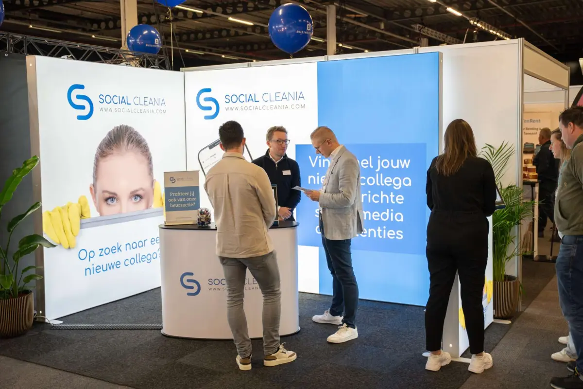 Beursstand van Social Cleania met blauw-witte wanden en het logo, waar bezoekers in gesprek zijn bij de balie